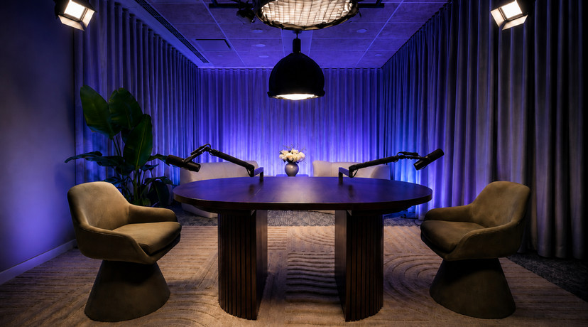 A modern podcast studio with two olive-green lounge chairs, a large round wooden table, multiple microphones, professional overhead lighting, floor-to-ceiling curtains, and a large indoor plant.