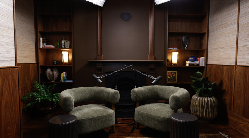 A warm, sophisticated podcast studio with two olive-green lounge chairs, microphones, wooden shelving, ambient lighting, plants, and a fireplace backdrop.