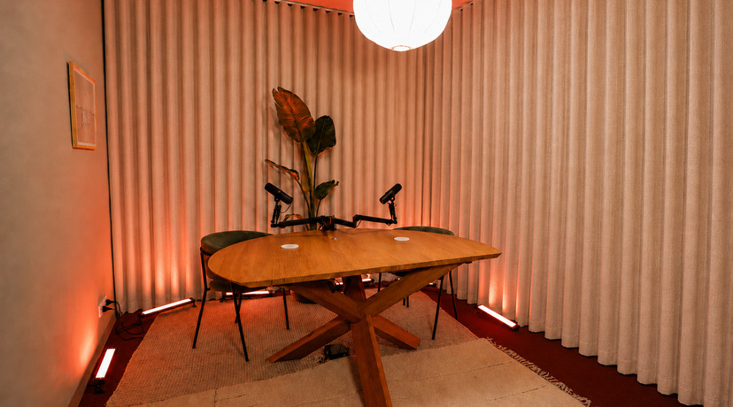 A warm, modern podcast studio featuring a large wooden table, two microphones, lounge chairs, indoor plants, cream curtains, and atmospheric orange lighting.
