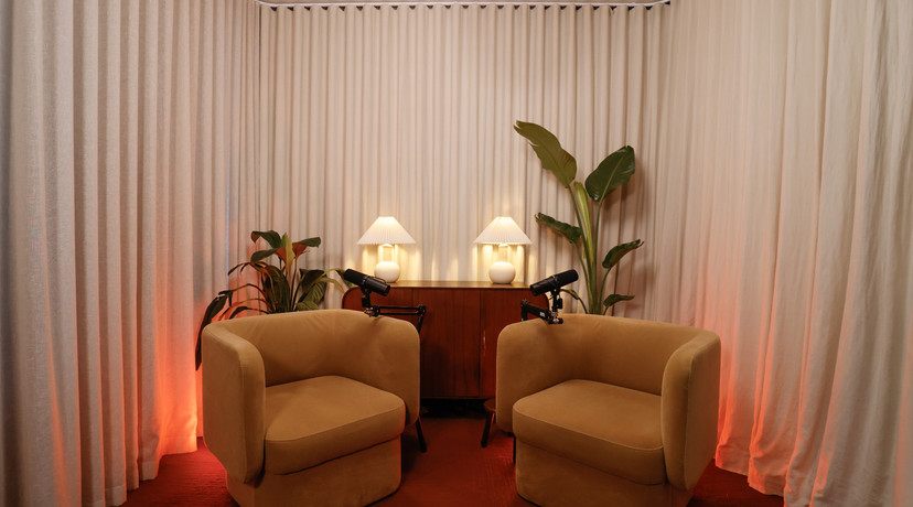 A warm, stylish podcast studio with two beige lounge chairs, microphones, indoor plants, soft table lamps, and cream curtains illuminated with orange accent lighting.