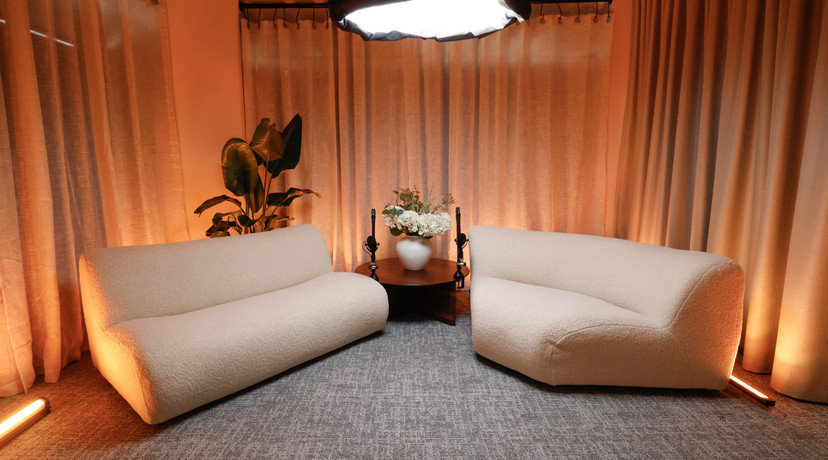 A stylish podcast studio with two cream boucle sofas, microphones, a small table with flowers, a large indoor plant, and warm orange accent lighting against soft curtains.